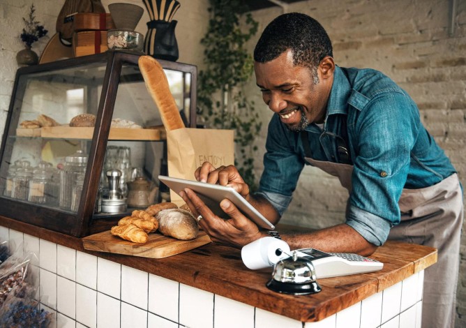 A smiling bakery employee is leaning over a counter and looking at his tablet.