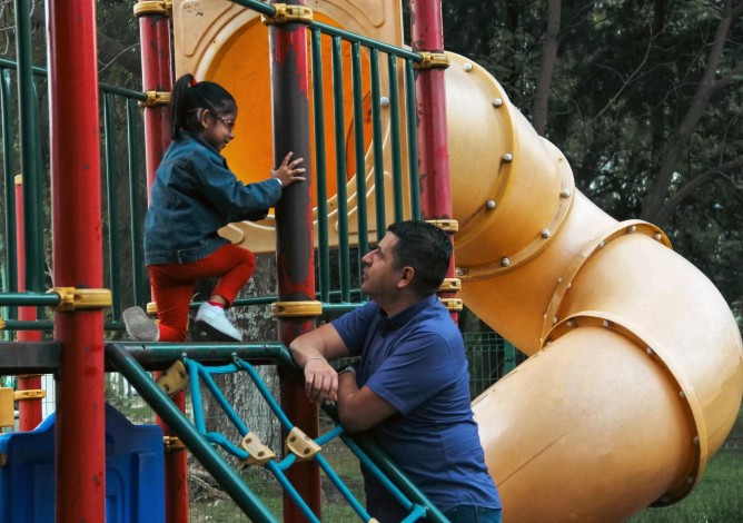 A young girl is on playground equipment that has a big slide; her father is leaning on the equipment looking at her.